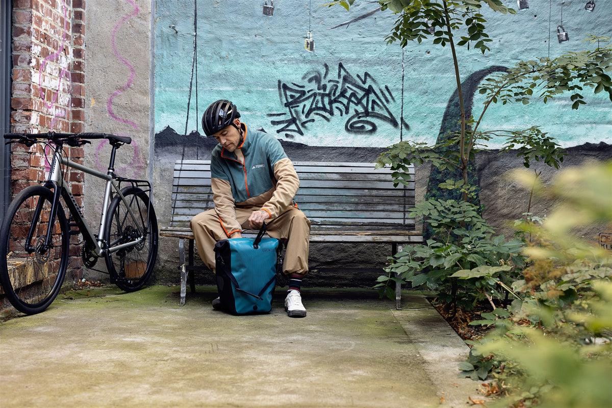 man wearing bike helmet opens ortlieb back-roller design bag in petrol blue on park bench